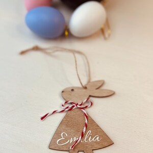 K&ouml;nnte beinhalten: Osterhase-Anh&auml;nger aus Holz mit dem Namen "Emilia" in wei&szlig;er Schrift. Der Hase hat eine rot-wei&szlig;e Schnur-Schleife und wird an einer nat&uuml;rlichen Schnur aufgeh&auml;ngt. Ostereier in Pastellfarben sind im Hintergrund.