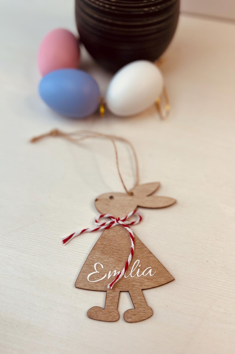 K&ouml;nnte beinhalten: Osterhase-Anh&auml;nger aus Holz mit dem Namen "Emilia" in wei&szlig;er Schrift. Der Hase hat eine rot-wei&szlig;e Schnur-Schleife und wird an einer nat&uuml;rlichen Schnur aufgeh&auml;ngt. Ostereier in Pastellfarben sind im Hintergrund.