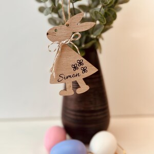 K&ouml;nnte beinhalten: H&ouml;lzerner Hasenanh&auml;nger mit einem dreieckigen Kleid, dem Namen "Simon" und Blumendetails. Der Hase h&auml;ngt an einer Schnur. Der Hintergrund umfasst eine Vase mit Gr&uuml;n und farbigen Ostereiern.
