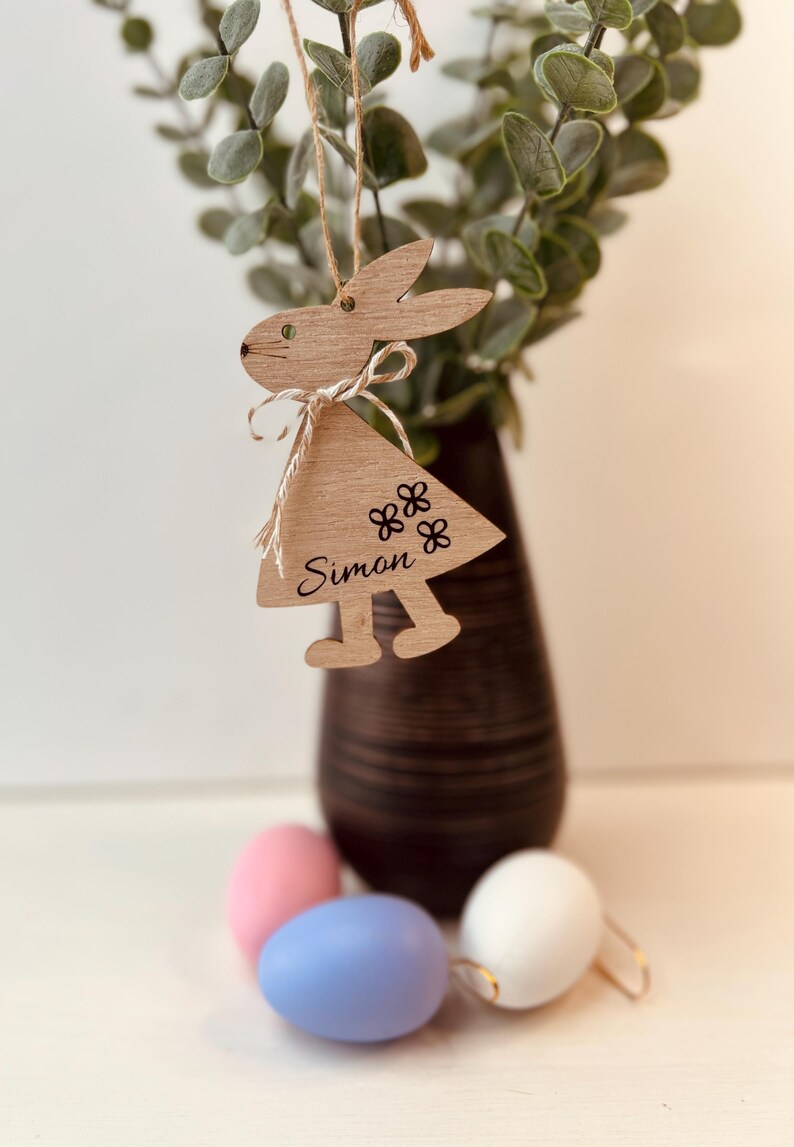 K&ouml;nnte beinhalten: H&ouml;lzerner Hasenanh&auml;nger mit einem dreieckigen Kleid, dem Namen "Simon" und Blumendetails. Der Hase h&auml;ngt an einer Schnur. Der Hintergrund umfasst eine Vase mit Gr&uuml;n und farbigen Ostereiern.