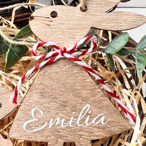 K&ouml;nnte beinhalten: Osterhase-Ornament aus Holz mit dem Namen "Emilia" in wei&szlig;er Schrift. Der Hase hat eine rot-wei&szlig;es Bindfaden-Schleife und ist von Stroh und Gr&uuml;n umgeben.