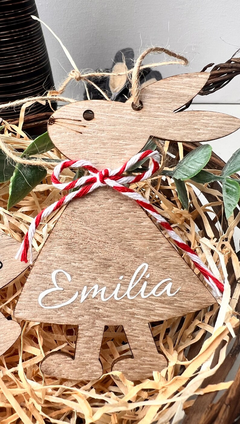 K&ouml;nnte beinhalten: Osterhase-Ornament aus Holz mit dem Namen "Emilia" in wei&szlig;er Schrift. Der Hase hat eine rot-wei&szlig;es Bindfaden-Schleife und ist von Stroh und Gr&uuml;n umgeben.