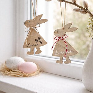 K&ouml;nnte beinhalten: Osterhasen-Dekorationen aus Holz, die an einem Zweig h&auml;ngen. Ein Hase tr&auml;gt den Namen "Simon" und Blumendetails, der andere den Namen "Emilia" und eine rot-wei&szlig; karierte Schleife. Zwei verzierte Eier liegen in einem Nest auf einer Fensterbank.