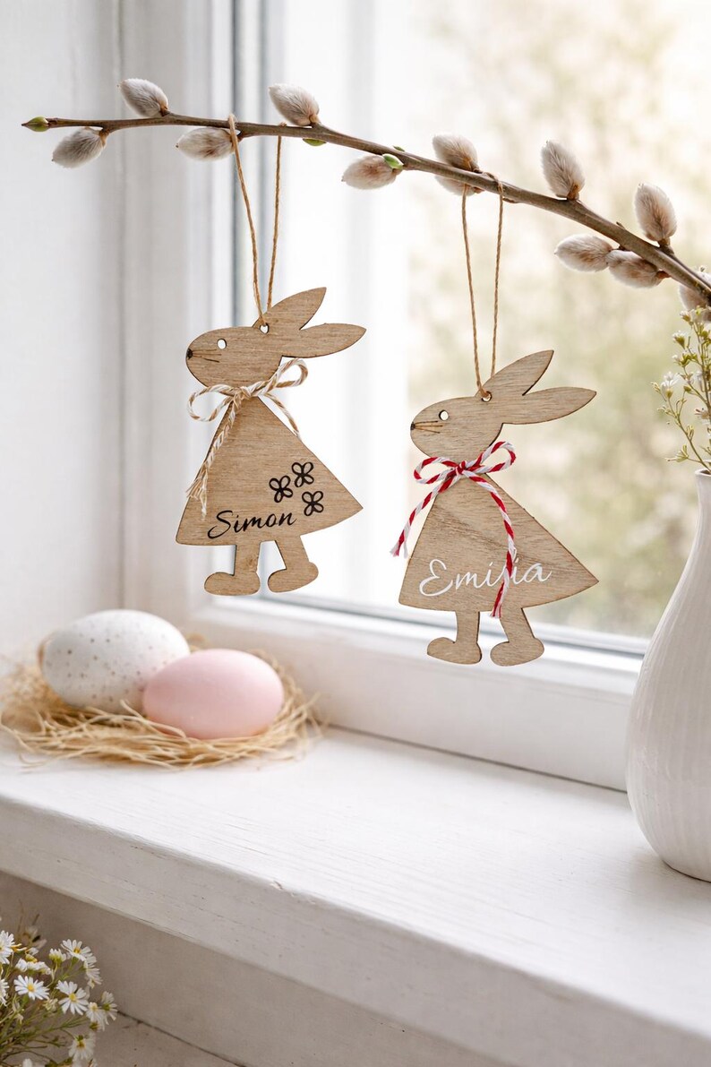 K&ouml;nnte beinhalten: Osterhasen-Dekorationen aus Holz, die an einem Zweig h&auml;ngen. Ein Hase tr&auml;gt den Namen "Simon" und Blumendetails, der andere den Namen "Emilia" und eine rot-wei&szlig; karierte Schleife. Zwei verzierte Eier liegen in einem Nest auf einer Fensterbank.