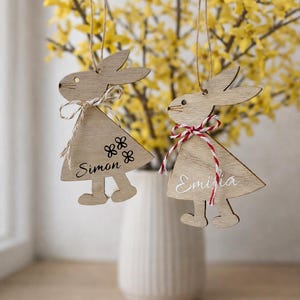 K&ouml;nnte beinhalten: Zwei h&ouml;lzerne Osterhasen-Anh&auml;nger mit den Namen "Simon" und "Emilia" h&auml;ngen vor gelben Bl&uuml;ten. Die Hasen haben Schleifen und sind aus hellem Holz gefertigt.