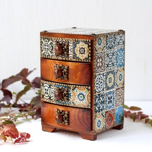 May include: A wooden box with four drawers, each decorated with a colorful tile pattern. The box has ornate metal handles and sits on a white surface.