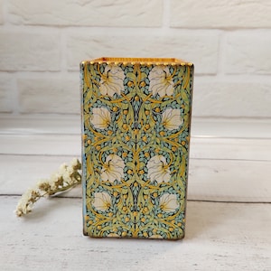 Può includere: Un portapenne in legno con un motivo floreale decorativo. Il motivo presenta fiori bianchi, foglie verdi e accenti dorati su uno sfondo blu.