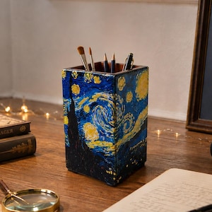 May include: A square wooden pencil holder featuring a hand-painted reproduction of The Starry Night with swirling blue, yellow, and black oil paint textures. The container holds several writing instruments and brushes, showcasing a deep, textured finish.