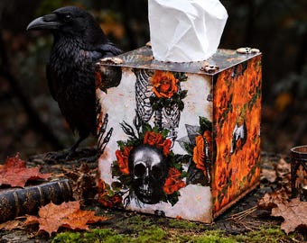 Puede incluir: Una funda cuadrada para caja de pañuelos con un estampado gráfico de un cráneo humano, una caja torácica esquelética y rosas de color naranja vibrante. La caja está construida con paneles planos y una abertura superior.