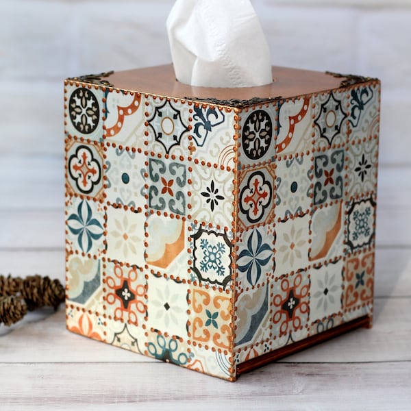 Puede incluir: Una funda cuadrada para caja de pañuelos con un patrón de azulejos multicolor en tonos naranja, azul y crema. La caja tiene bordes metálicos en tono cobre y detalles decorativos en las esquinas, con un pañuelo blanco saliendo por arriba.