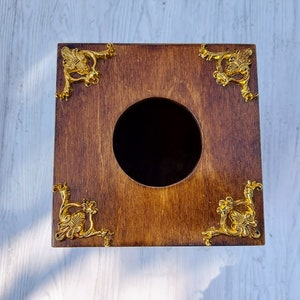 May include: A square wooden tissue box cover with a dark brown stained finish. The top surface features a central circular opening and four ornate, metallic gold-colored decorative corner accents with intricate scrollwork and floral-like patterns.