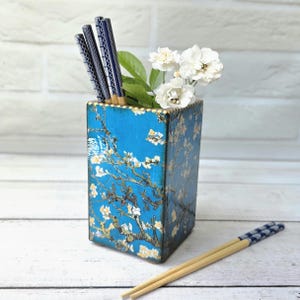 May include: A rectangular blue ceramic holder decorated with a white almond blossom pattern, containing three dark blue patterned chopsticks and a small bouquet of white flowers, with two additional chopsticks lying on the surface.