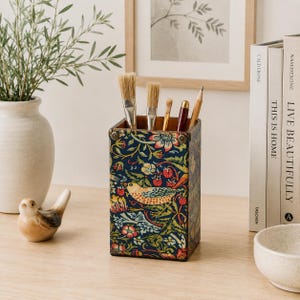 May include: A rectangular pencil holder featuring a dark blue background with a colourful botanical pattern of birds, strawberries, and flowers. The top corners are accented with small metallic studs. It contains several paintbrushes and a pencil.