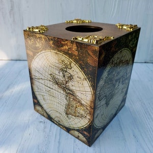 May include: A square tissue box cover featuring a vintage world map print in shades of brown and cream. The top surface includes a circular opening and four decorative gold-toned metal corner accents with an ornate, raised relief design.