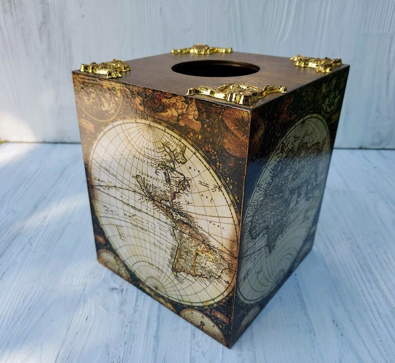 May include: A square tissue box cover featuring a vintage world map print in shades of brown and cream. The top surface includes a circular opening and four decorative gold-toned metal corner accents with an ornate, raised relief design.
