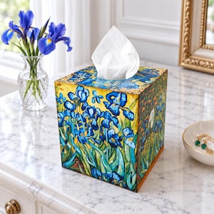 May include: A square tissue box cover featuring a vibrant print of blue iris flowers with green stems against a yellow background. The box is topped with a white tissue and has gold-toned decorative metal corners on the top edges.