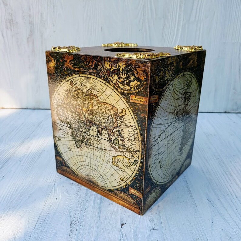 May include: A square tissue box cover featuring a vintage world map print in shades of brown, cream, and black. The top surface includes a circular opening and four decorative gold-toned metal corner accents with intricate embossed detailing.