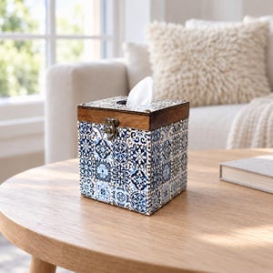 May include: A square tissue box cover featuring a blue and white patterned tile design on the sides. The top is made of dark brown wood with a metal latch closure on the front. A single white tissue is visible protruding from the top opening.