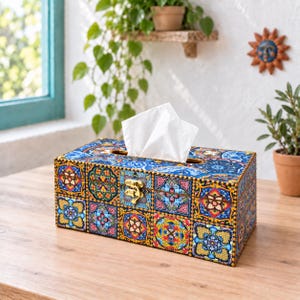 May include: A rectangular wooden tissue box cover featuring a vibrant, multi-coloured tile pattern in shades of blue, yellow, red, and green. The box has a hinged lid with a gold-toned metal latch and holds a stack of white facial tissues.