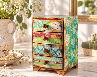 May include: A small wooden chest of drawers featuring four stacked compartments. Each drawer front displays a unique, colorful floral and damask pattern in shades of teal, green, red, and gold. The unit stands on four short feet and has round knobs.