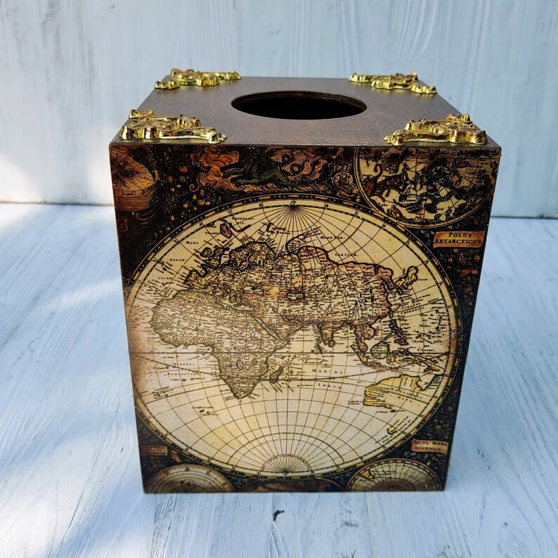 May include: A square tissue box cover featuring a vintage-style world map print in sepia and cream tones. The top surface has a circular opening and is accented with ornate, metallic gold-colored corner brackets on each of the four corners.