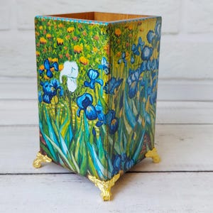 Puede incluir: Un portalápices rectangular de madera con un diseño floral con iris azules, flores amarillas y hojas verdes. El soporte tiene patas doradas. El estilo artístico recuerda a una pintura.