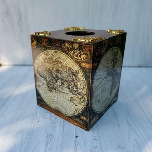May include: A square tissue box cover featuring a vintage-style world map print in shades of brown, cream, and black. The top surface has a circular opening and is decorated with ornate, metallic gold-colored corner accents on each edge.