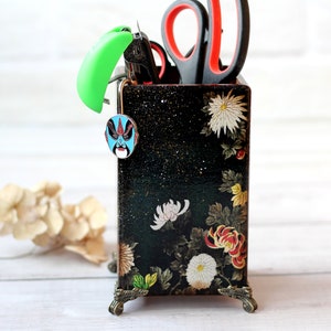 Peut inclure: Un organisateur de bureau rectangulaire noir orné d'un motif floral de chrysanthèmes blancs et rouges. Le récipient possède des pieds en métal bronze et contient des ciseaux, un outil vert vif et une petite broche masque d'opéra bleu.