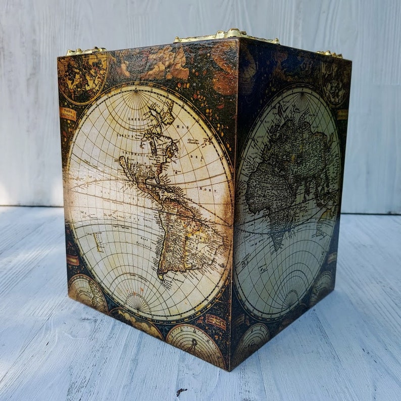 May include: A square wooden box featuring a vintage-style world map print on its sides. The map displays continents in sepia and cream tones against a dark, textured background. Gold-colored metal hardware accents the top corners of the box.