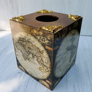 May include: A square wooden tissue box cover featuring a vintage world map print in sepia and cream tones. The top surface has a circular opening and is decorated with four ornate, metallic gold-colored corner accents in a filigree style.