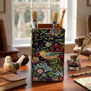 May include: A rectangular wooden pencil holder featuring a dark blue background with a colourful pattern of birds, strawberries, and green foliage. The holder contains three paintbrushes and is decorated with small metal studs at the top corners.