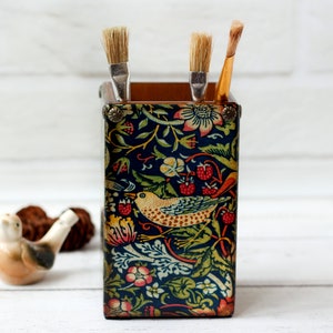 Puede incluir: Un porta lápices de cerámica con estampado floral azul y verde con tres pinceles que sobresalen de la parte superior. El porta lápices presenta un diseño de pájaro con bayas rojas.