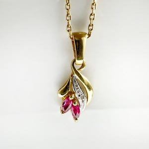 Op de afbeelding: Een gouden hanger met twee marquise-geslepen rode robijnen en een zilverkleurig accent bezet met kleine heldere steentjes. De hanger hangt aan een fijne gouden ketting en heeft een gebogen, bladvormig ontwerp van gepolijst geelgoud.