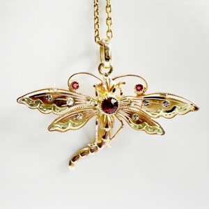 May include: A gold dragonfly pendant necklace featuring a central round dark red gemstone on the thorax, two smaller red stones on the upper wings, and several clear white accent stones embedded along the textured, scalloped wing edges.