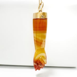 Puede incluir: Un colgante con un puño tallado en ágata bandeada naranja y amarilla. El colgante cuelga de una cadena de oro y tiene una tapa de oro con el número "750" estampado.