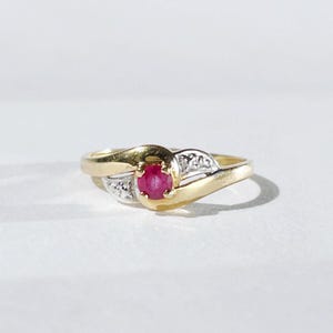Op de afbeelding: Een gouden ring met een centrale, ronde, rode edelsteen. De ring heeft een split-schachtontwerp met kleine, heldere accentstenen aan weerszijden van de centrale steen. De ring is geplaatst tegen een witte achtergrond.