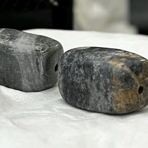 May include: Two dark gray, polished stone beads with a hole drilled through each. The beads have a smooth, rounded shape and a mottled appearance, with variations in color from light to dark gray. They rest on a white surface.