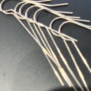 May include: A collection of silver-colored earring hooks, displayed against a dark background. The hooks have a simple, curved design with a long, straight dangle. The earrings are likely for pierced ears.