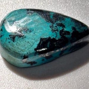 Puede incluir: Una gema pulida en forma de lágrima con un patrón llamativo. La piedra presenta un vibrante color base turquesa, contrastado por inclusiones negras irregulares. La superficie es lisa y reflectante, mostrando variaciones naturales.