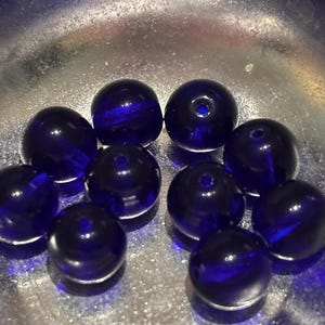 Puede incluir: Una colección de diez cuentas de vidrio azul oscuro, cada una esférica con un pequeño agujero. Las cuentas se muestran en un cuenco plateado, que refleja la luz. Las cuentas tienen un acabado brillante y parecen hechas a mano.