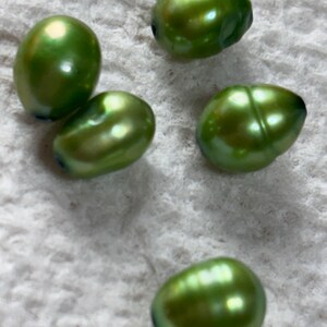 May include: Five lustrous, green-hued pearls with an iridescent sheen. The pearls have an oval shape and are scattered on a textured, off-white surface. The pearls reflect light, showcasing their natural beauty and unique color.