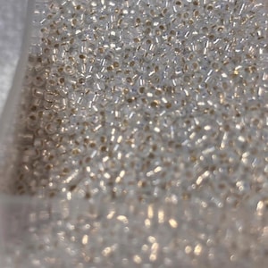 May include: A close-up shot of a clear plastic container filled with small, clear, cylindrical beads. The beads have a slight golden hue in the center. The text "G-C-R-7g-273" is visible in the upper left corner. These beads are suitable for jewelry making and crafts.