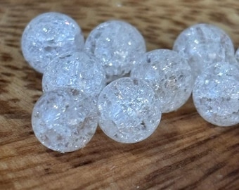 Può includere: Primo piano di diverse perline di vetro trasparente con effetto craquelé. Le perline sferiche hanno un aspetto strutturato e fratturato. Sono disposte su una superficie di legno, creando un contrasto tra le perline lisce e le venature del legno. Ogni perlina misura circa 1 cm di diametro.