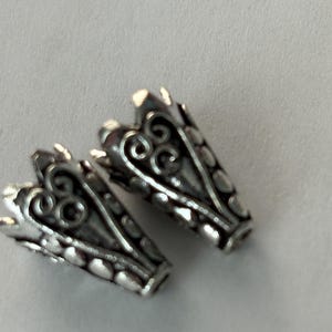 Könnte beinhalten: Zwei silberfarbene Metallperlenkappen mit einem verzierten Filigran-Design. Jede Kappe hat eine zylindrische Form mit einem dekorativen, gewellten Rand und aufwendigen Wirbelmustern. Die Kappen werden vor einem schlichten, hellfarbigen Hintergrund gezeigt.