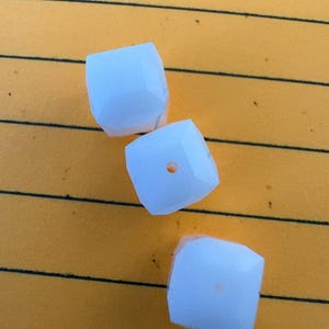 May include: Three white, faceted, cube-shaped beads arranged in a diagonal line. Each bead features a smooth, geometric surface with a small, centered hole for threading, showcasing a clean, opaque appearance and uniform, angular edges.