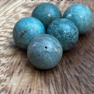 Puede incluir: Cinco cuentas esféricas de color turquesa con diferentes texturas y patrones. Una cuenta tiene un pequeño agujero. Las cuentas están dispuestas sobre una superficie de madera. El texto "N-G-R-12-05-207" es visible en la parte superior. Estas cuentas son adecuadas para la fabricación de joyas.