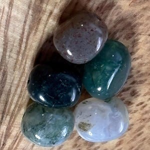May include: A collection of six polished gemstones in various colors, including green, blue, and purple. The stones are smooth and rounded, with natural patterns and textures. They are displayed on a wooden surface.