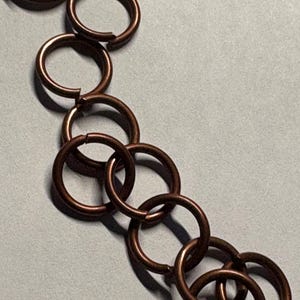 May include: A collection of circular copper-colored jump rings arranged in a loose, overlapping chain pattern. Each individual ring features a small, visible split or opening along its circumference, showing a metallic finish and smooth texture.