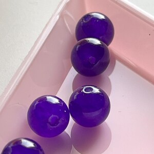 May include: Close-up of six deep purple, spherical beads with a glossy finish, resting in a light pink rectangular container. The beads have a smooth surface and are evenly spaced within the container.
