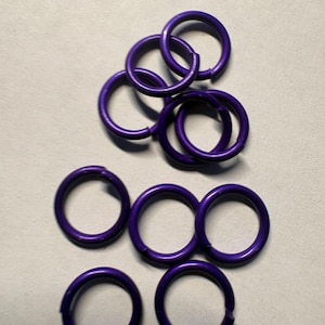May include: A collection of nine small, circular purple jump rings, each featuring a single cut or opening along the circumference. These metal rings are arranged in a loose cluster, displaying a consistent smooth, matte purple finish.